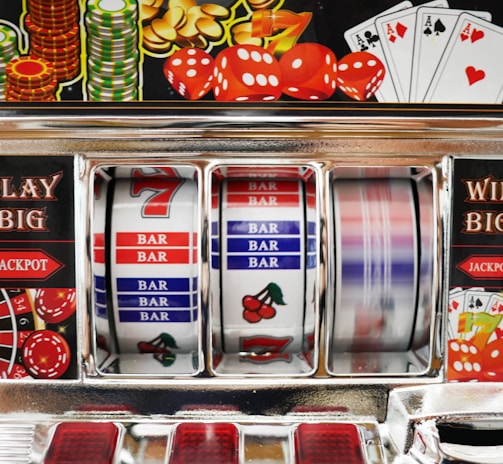 A close-up view of a slot machine with vibrant colors and various gambling symbols such as dice, coins, and playing cards. The reels display a combination of symbols, including bars, cherries, and the number seven. The chrome exterior and colorful details convey the theme of casino and gambling.