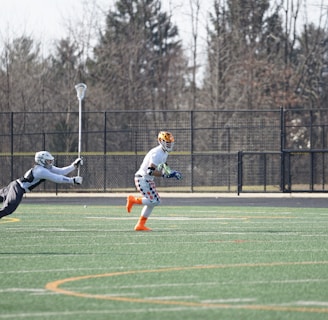 A group of athletes engaged in a sports game on a green field. One player is running forward with possession of the ball, wearing colorful socks and a checkered uniform, while another player in dark clothing dives to intercept or tackle. Other players are present in the background. Leafless trees and a metal fence surround the field.