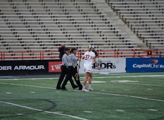 A player in a white sports uniform with the number 37 is standing on a sports field, accompanied by two referees in black and white striped shirts. The background shows empty stadium seating and advertising banners along the sidelines.