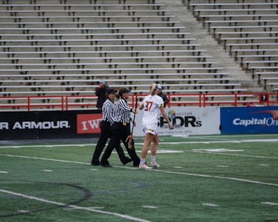 A player in a white sports uniform with the number 37 is standing on a sports field, accompanied by two referees in black and white striped shirts. The background shows empty stadium seating and advertising banners along the sidelines.