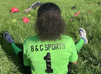 A person with curly hair sits on green grass facing away, wearing a bright green sports jersey with 'B & G SPORTS' and the number 1 on the back. Sports equipment, including a red helmet and baseballs, is scattered around.