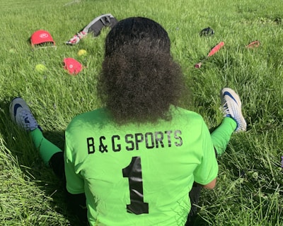 A person with curly hair sits on green grass facing away, wearing a bright green sports jersey with 'B & G SPORTS' and the number 1 on the back. Sports equipment, including a red helmet and baseballs, is scattered around.