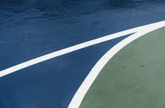 A close-up view of a sports court featuring distinctly marked lines, with a dark blue surface contrasted by a curve of green. The white lines delineate the boundary between the two areas, creating an abstract visual effect.