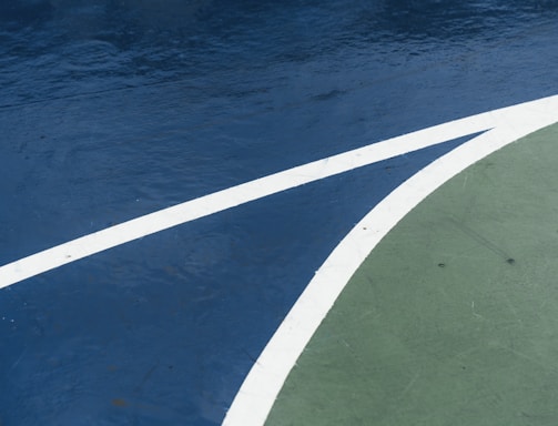 A close-up view of a sports court featuring distinctly marked lines, with a dark blue surface contrasted by a curve of green. The white lines delineate the boundary between the two areas, creating an abstract visual effect.