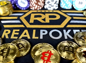 Stacks of poker chips in various colors are arranged in the background, while in the foreground, there are numerous golden coins featuring the Bitcoin symbol. The text 'REALPOKER' is prominently displayed in gold and silver lettering on a dark background.