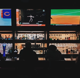 A bar counter with several people sitting in front of it is shown. Above the counter, there are multiple television screens displaying sports broadcasts, including football and baseball. Behind the counter, various bottles of alcohol are neatly arranged.