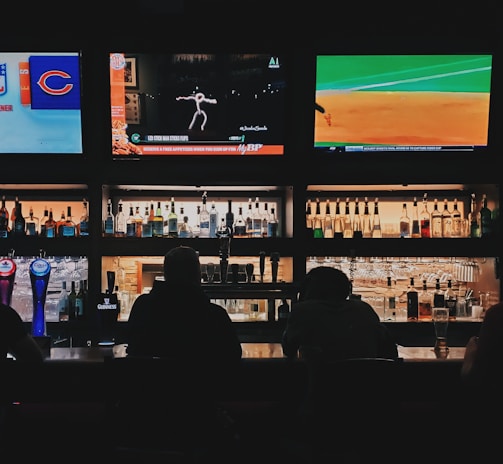 A bar counter with several people sitting in front of it is shown. Above the counter, there are multiple television screens displaying sports broadcasts, including football and baseball. Behind the counter, various bottles of alcohol are neatly arranged.