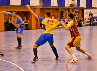 A dynamic indoor sports scene with players in motion on a court. Two athletes in blue and yellow uniforms are engaged in an intense handball game, with one player gripping the other's jersey. The background shows other players, several seated people, and advertisements on the walls.
