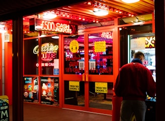 A brightly lit entrance to a casino with neon signs, including one advertising a £500 jackpot. The doors have clear signage indicating entry is restricted to those over 18. A person in a red jacket stands to the right, possibly interacting with a machine or display.
