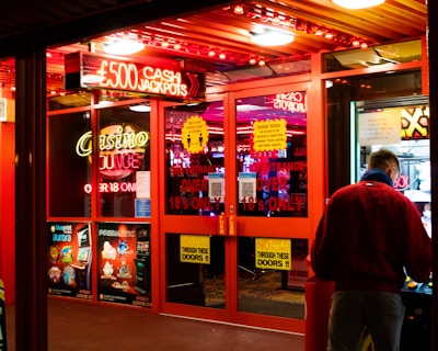 A brightly lit entrance to a casino with neon signs, including one advertising a £500 jackpot. The doors have clear signage indicating entry is restricted to those over 18. A person in a red jacket stands to the right, possibly interacting with a machine or display.