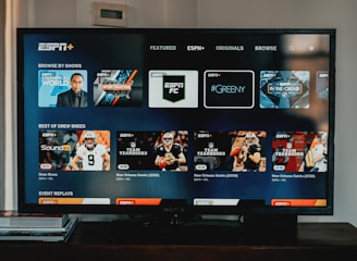 A television screen displaying the ESPN+ streaming interface with various sports-related shows and categories. The screen includes sections such as 'Browse by Shows' and 'Best of Drew Brees' with thumbnails of sports programs featuring athletes in action and analysts.