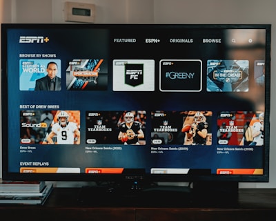 A television screen displaying the ESPN+ streaming interface with various sports-related shows and categories. The screen includes sections such as 'Browse by Shows' and 'Best of Drew Brees' with thumbnails of sports programs featuring athletes in action and analysts.