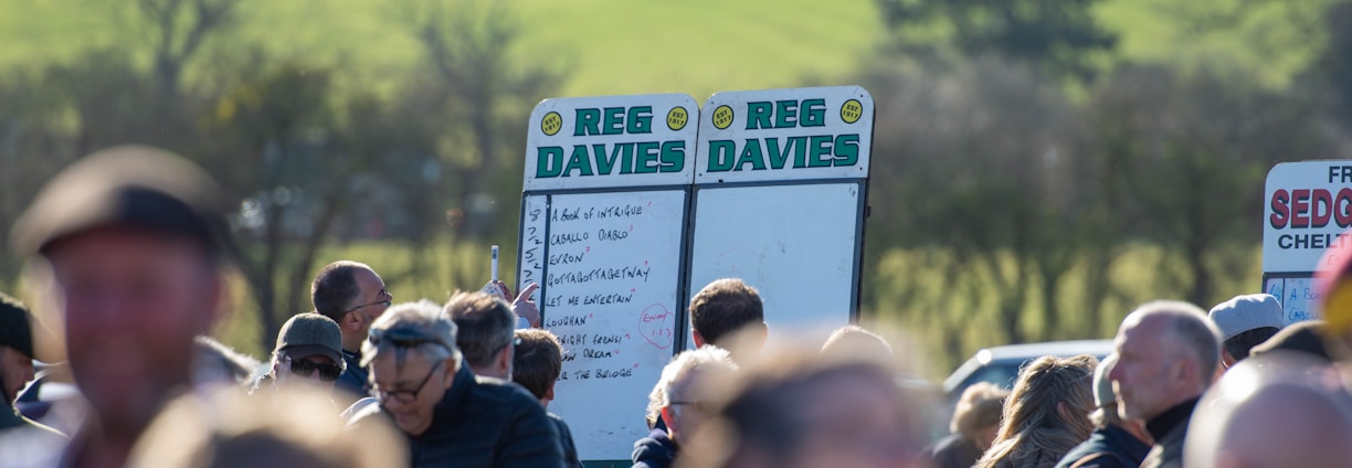 A group of people gathered outdoors with two large betting boards in the background labeled 'REG DAVIES'. The setting appears to be a countryside location with trees and fields visible in the distance. The crowd consists of individuals dressed in jackets and hats, engaging in conversation.