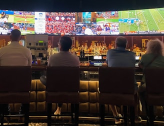 A group of people sit at a bar with their backs to the camera, watching multiple sports games displayed on large screens. The bar counter is lined with various bottles of liquor, and the atmosphere suggests a casual and relaxed social setting.