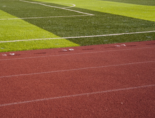 A sports field with vivid green grass divided into sections by white lines in the background. In the foreground, a track with a reddish-brown surface is marked with numbers and lines.