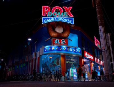 A vibrant, neon-lit facade of a gaming and sports establishment at night, featuring a large glove holding a baseball above the entrance. The building is adorned with colorful signs in both English and Japanese, with various advertisements and an illuminated sign that says 'ROX DOME'. Several people are walking towards the entrance, and bicycles are parked along the side.