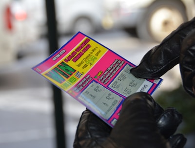 A person wearing black gloves is holding a colorful lottery scratch-off ticket. The ticket includes information about a $1,000,000 guaranteed minimum payout and features multiple games with various prize amounts.