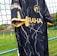 A black sports jersey with a marbled white pattern hangs on a net fence. The jersey features yellow accents on the sleeves and collar, and logos in yellow on the front and side. The background includes a blurred, grassy field.